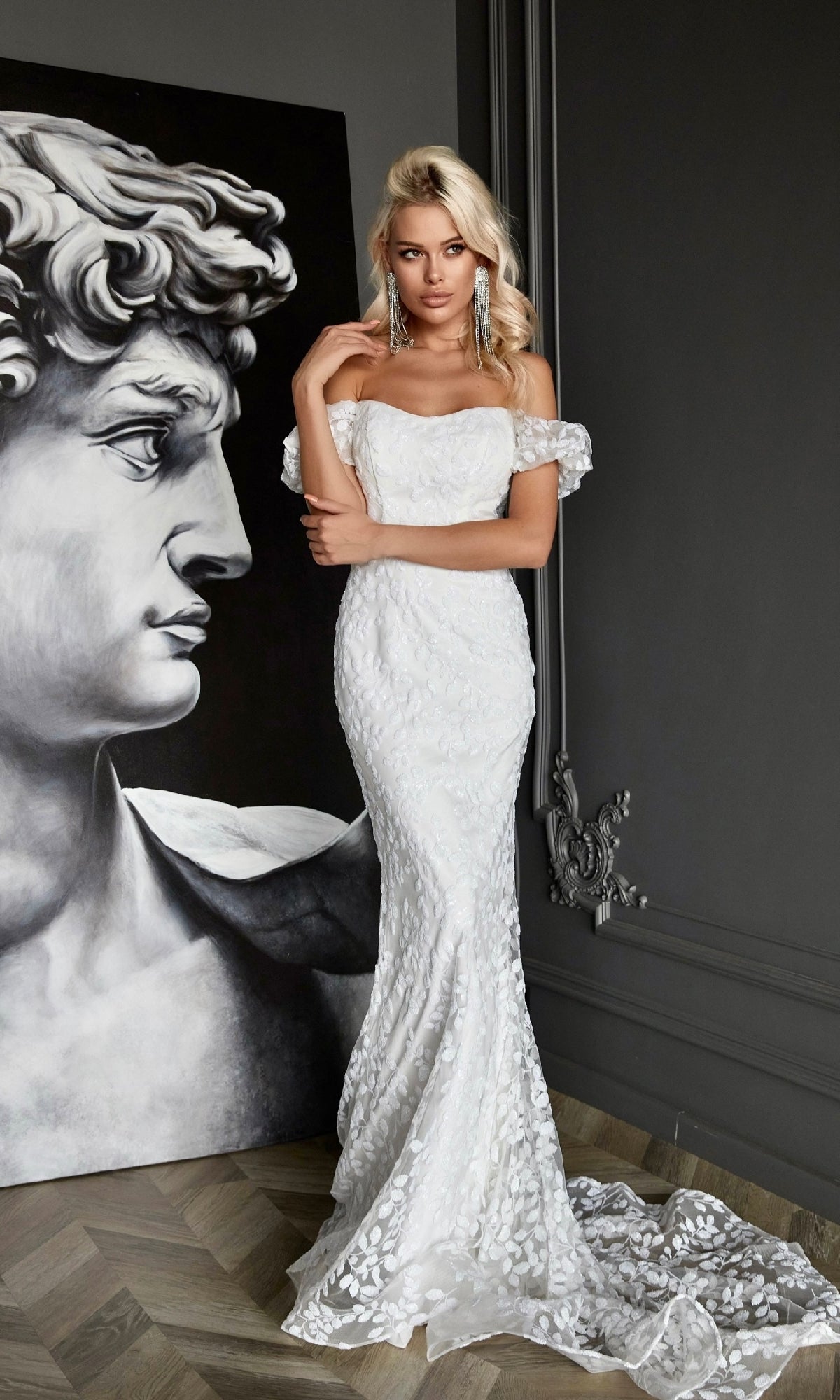 A woman wearing the Off-Shoulder White Formal Dress: Tina Holly Tk024W, a floor-length lace evening gown, stands beside a large grayscale painting of a classical statue head in an elegant interior.